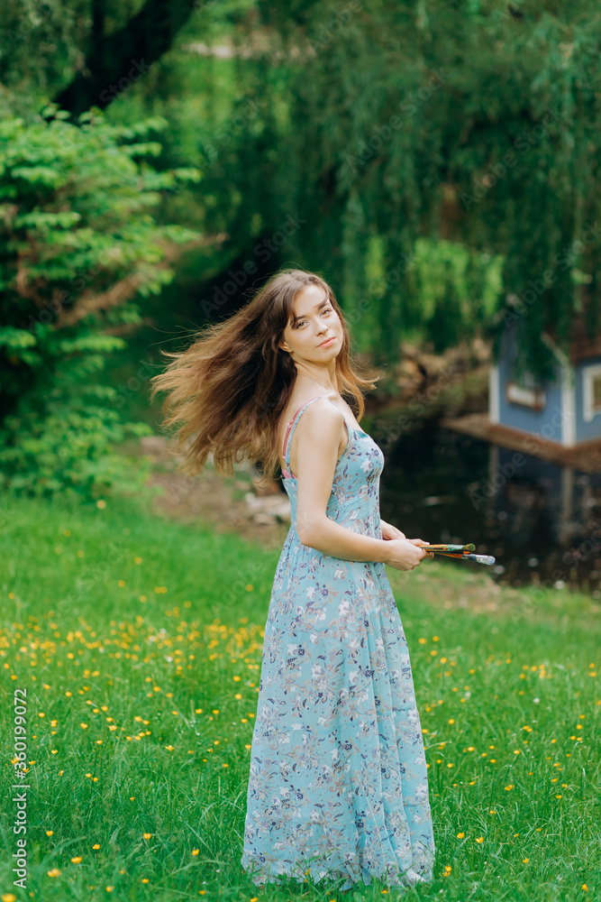 Beautiful girl in blue dress stands near an easel holding a brush. Girl painter artist is painting outdoors. Beautiful girl in blue dress stands near an easel holding a brush. Girl painter artist is painting outdoors.