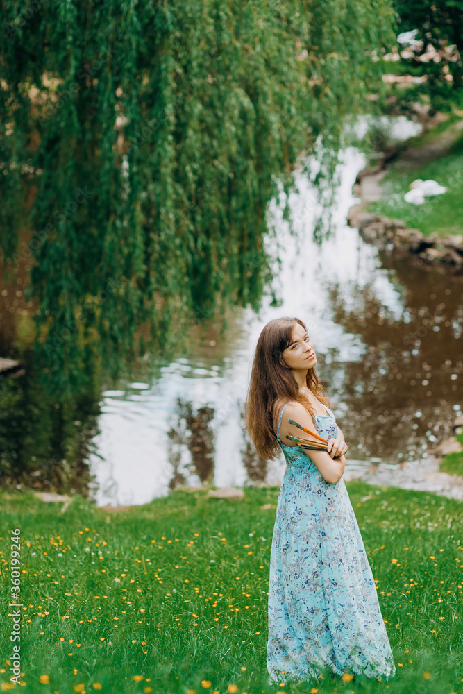 Beautiful girl in blue dress stands near an easel holding a brush. Girl painter artist is painting outdoors. Beautiful girl in blue dress stands near an easel holding a brush. Girl painter artist is painting outdoors.