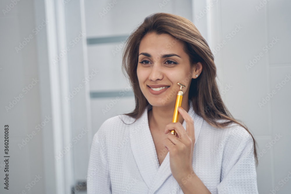 Cheerful young woman using golden facial massager Cheerful young woman using golden facial massager