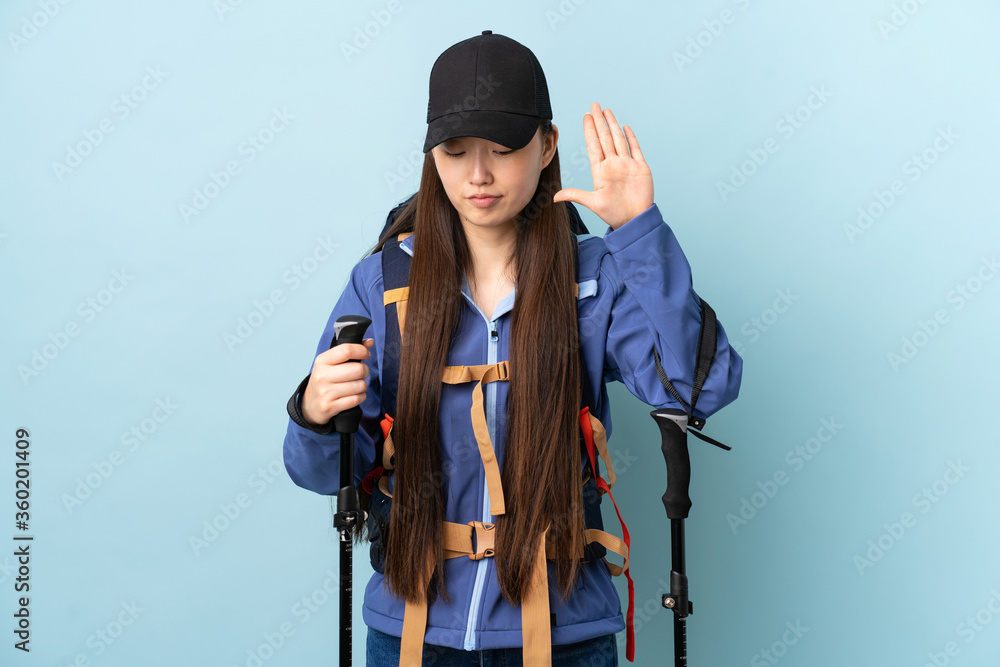 Young Chinese girl with backpack and trekking poles over isolated blue background making stop gesture and disappointed Young Chinese girl with backpack and trekking poles over isolated blue background making stop gesture and disappointed
