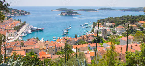 View from fortress of Hvar Harbor below with marina filled with sailboats and yachts, orange rooftops of village below and other Croatian Islands in the Adriatic in the distance on a clear summer day.