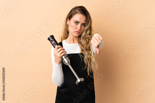 Young brazilian woman using hand blender isolated on beige background showing thumb down with negative expression