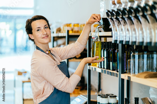 Young saleswoman in apron, protective mask and gloves pouring oil in glass bank or jar in zero waste shop. Female owner selling superfoods in zero waste shop. Reopening after coronavirus pandemic.