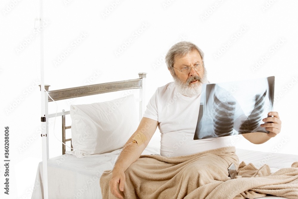 Elderly old man recovering in a hospital bed isolated on white background. Getting care. Concept of healthcare and medicine. Close up of hand with IV for treatment, looking on lung scan. Copyspace. Elderly old man recovering in a hospital bed isolated on white background. Getting care. Concept of healthcare and medicine. Close up of hand with IV for treatment, looking on lung scan. Copyspace.