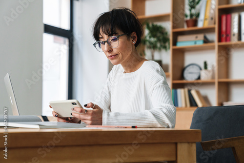 Photo of focused woman using smartphone while working with laptop