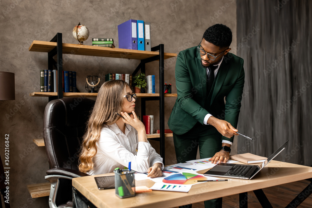 Work in a creative office. An African man and a beautiful business lady in formal clothes are working together, standing indoors Work in a creative office. An African man and a beautiful business lady in formal clothes are working together, standing indoors