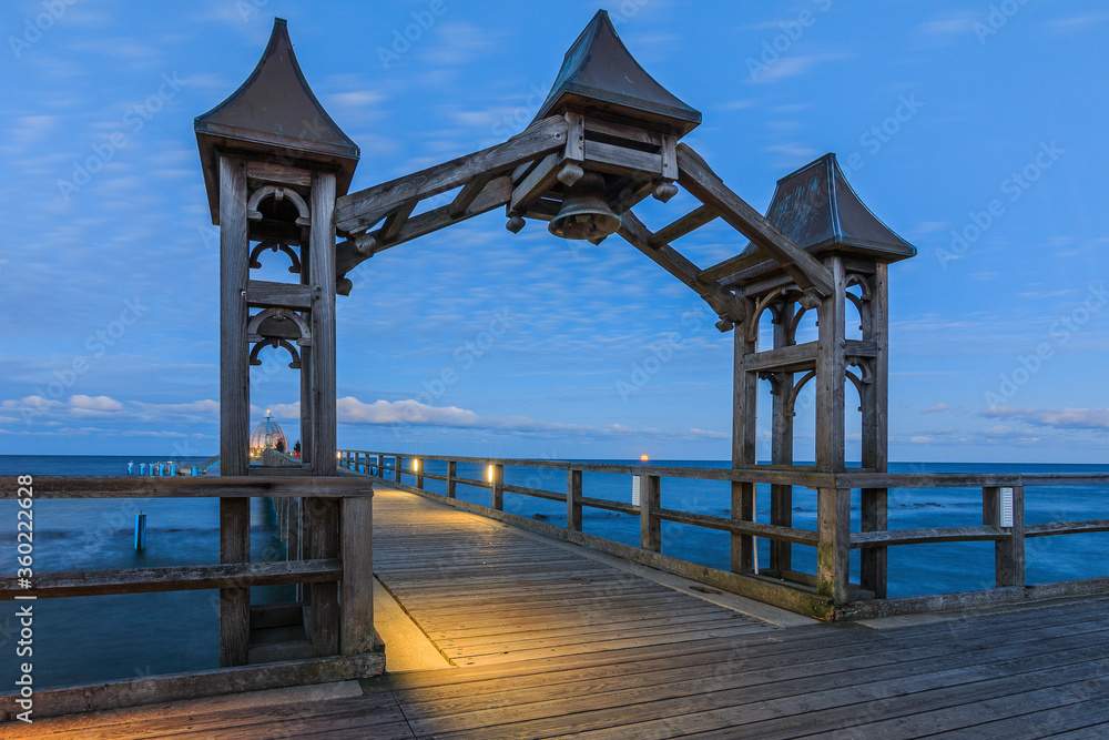 Wooden pier on the Baltic Sea in the evening. Lateral view of the sunset with clouds on the horizon. Illuminated footbridge with wooden arch and bell on the island of Ruegen