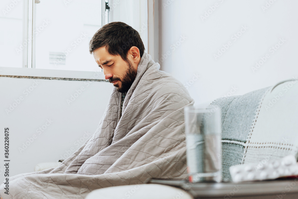 Obraz premium Man feeling sick on the couch at home with cold and flu symptoms. Medicine and glass of water in the foreground.
