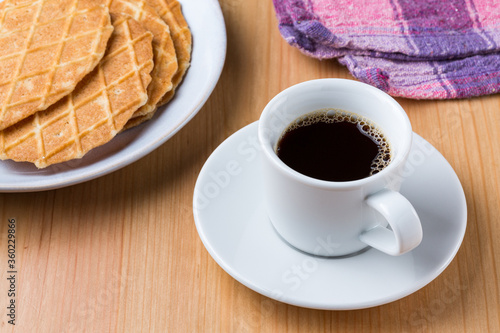 A cup of dark hot black coffee and pile of waffle biscuits on a white plate dish on a wood table