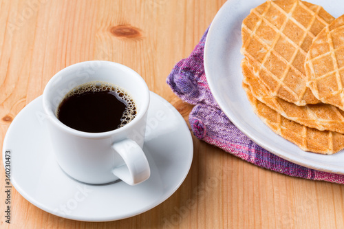 A cup os coffee and a pile of waffle biscuits in a plate on a wood table