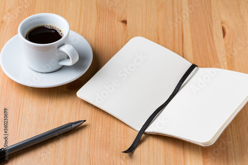 A light wood desktop table with a cup of coffee, a pen and a open notebook
