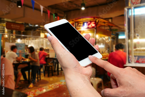 Close up hands hold and touch screen smartphone with blank screen for put your own webpage or message over blurred food court background. Ordering food online, online shopping, new normal concept