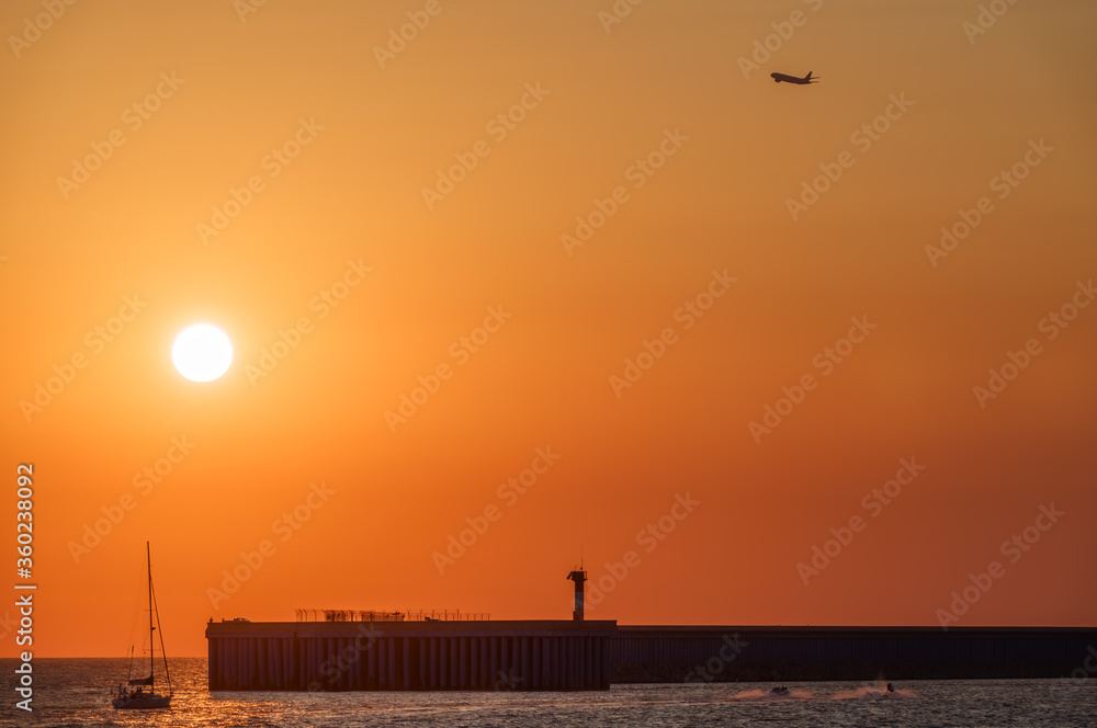 Beautiful red and orange sunset over the sea. The sun goes down over the sea. Silhouette of a take-off airplane and the setting sun. Silhouette of sea port at sunset