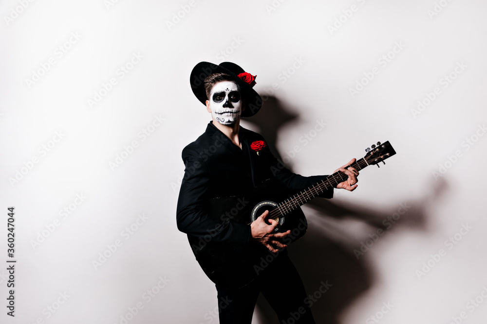 Studio portrait of mexican mariachi in deadman attire. Indoor photo of male model with zombie makeup playing guitar. Studio portrait of mexican mariachi in deadman attire. Indoor photo of male model with zombie makeup playing guitar.