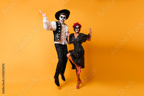 Glad zombie man in sombrero jumping on yellow background. Charming muerte girl in black dress dancing with boyfriend.