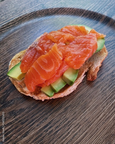 tasty fat aromatic sandwich with white fresh bread, croutons, avocado and salmon lies on a dark round wooden plate and stands on the table, top view