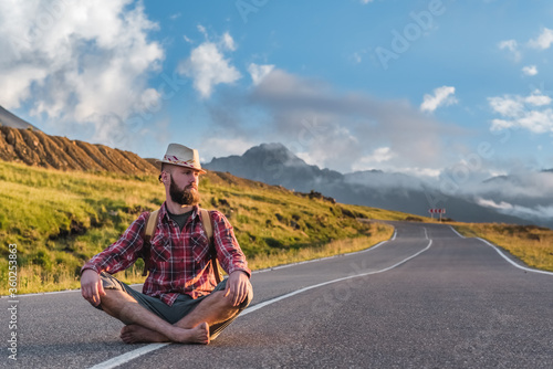 Handsome young man, in a plaid shirt and shorts, sits on a track in the mountains at sunset. Freedom and travel hitchhiking concept