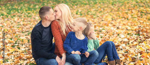 Portrait of young family sitting in autumn leaves. Parents kissing and sitting with children in the autumn park