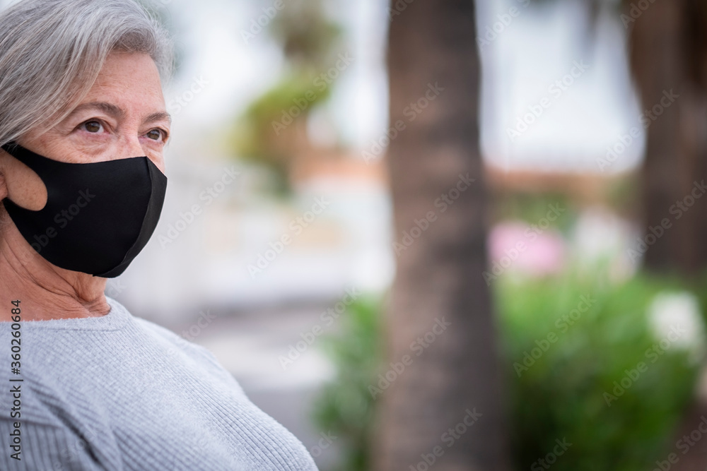 Pensive white-haired senior woman with medical mask due to coronavirus sitting in a public park with tall palm trees - love of nature and new normal life Pensive white-haired senior woman with medical mask due to coronavirus sitting in a public park with tall palm trees - love of nature and new normal life
