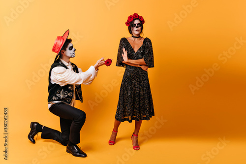 Latin man in love standing on knee beside his girlfriend. Cheerful zombie guy with rose posing on yellow background with brunette girl.