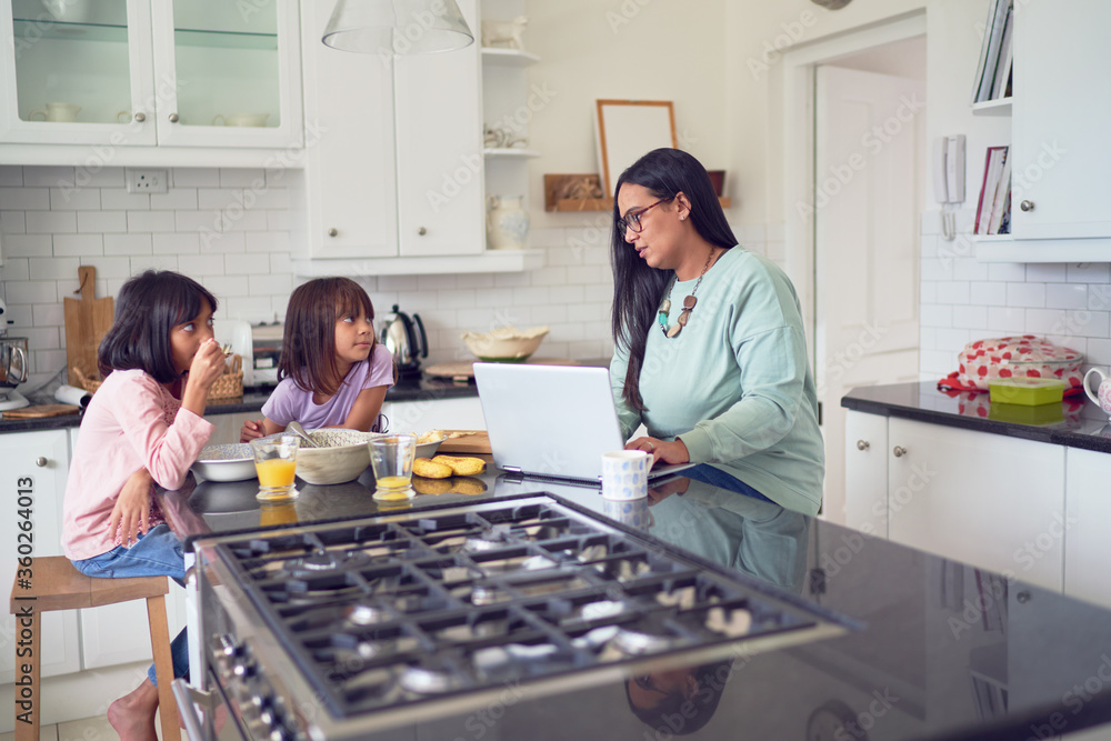 Mother working at laptop while daughters eat breakfast in kitchen Mother working at laptop while daughters eat breakfast in kitchen
