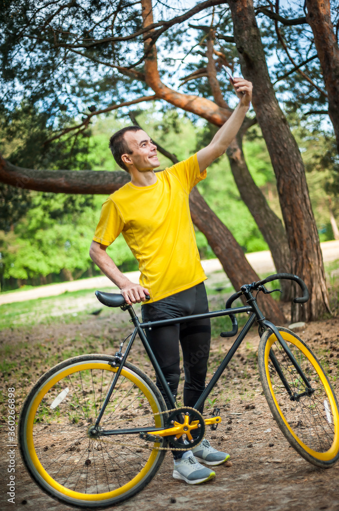 Naklejka premium A young man taking a selfie with his Bicycle. In a public Park, among trees and vegetation.