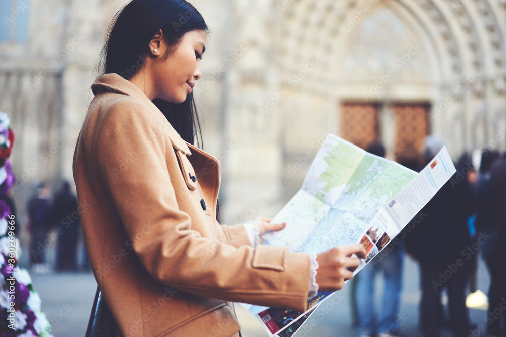 Attractive young female hipster in trendy spring coat fond of traveling spent short weekends in old european town using map to find tourist route to get to popular notable places in downtown - obrazy, fototapety, plakaty 