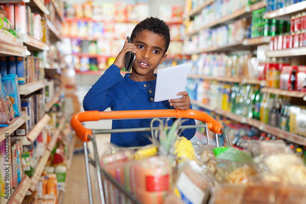Focused African tween boy talking on phone and looking at shopping list while visiting supermarket