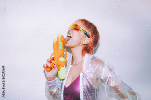 Stylish young female in retro futuristic outfit licking water pistol with closed eyes against gray background photo