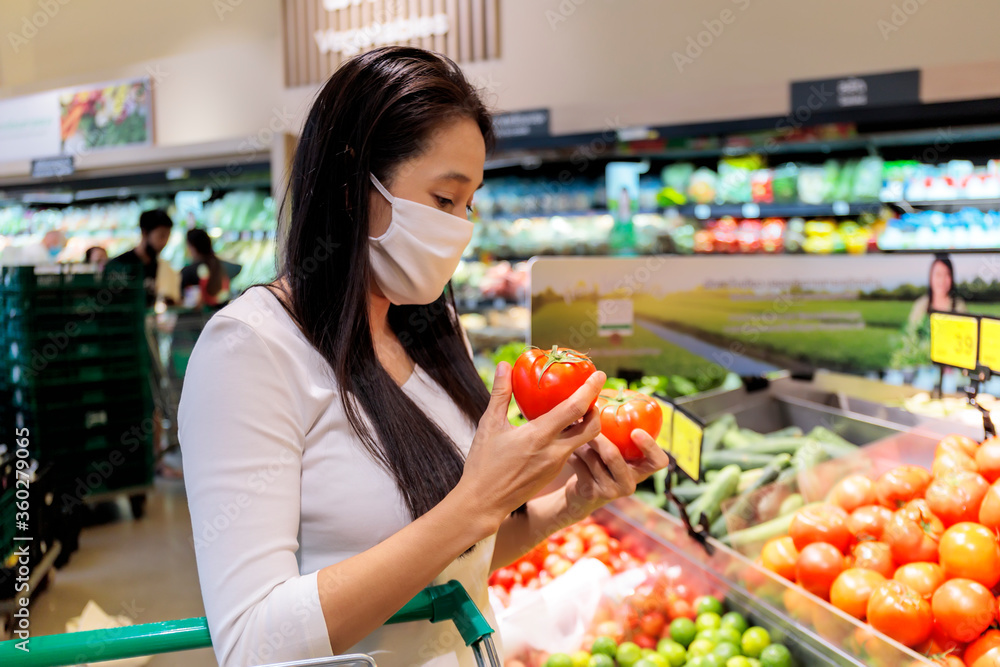 Asian woman wearing protective face mask push and hold shopping cart in supermarket department store. Girl, looking grocery to buy  some food. New normal after covid-19. Family concept.