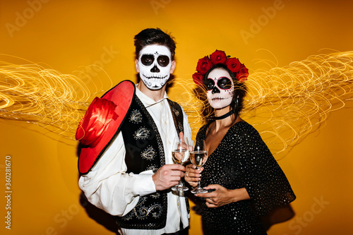 Zombie man in red sombrero drinking wine. Studio portrait of happy dead couple posing on golden background in halloween.