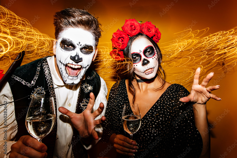 Laughing zombie guy drinking champagne at party with friend. Amazing girl in muertos costume enjoying wine with boyfriend. Laughing zombie guy drinking champagne at party with friend. Amazing girl in muertos costume enjoying wine with boyfriend.