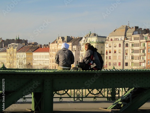 Budapest, Hungary - March 16, 2020: Young people sitting on the green Freedom bridge side in front of city panorama in the empty city due to coronavirus covid-19 in Budapest, Hungary