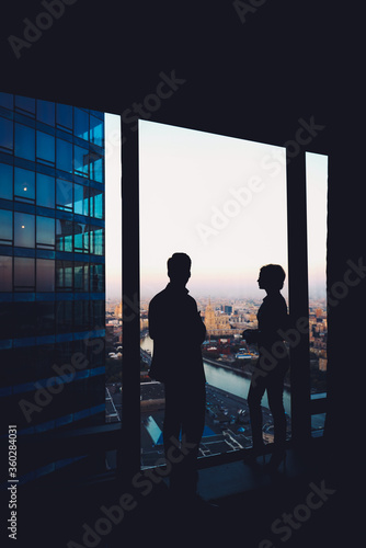 Silhouette of two confident colleagues having conversation while standing in corridor big company near window in evening time, young skilled workers discuss ideas after business meeting with partners