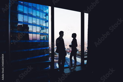 Silhouette of a couple of young business people having conversation while standing in hallway company near window, intelligent successful man and woman office workers discuss ideas during break