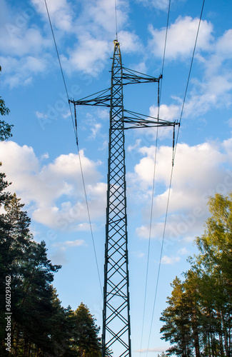 High voltage metal tower electricity pole and power lines on the blue sky background with white clouds on a sunny day in the forest