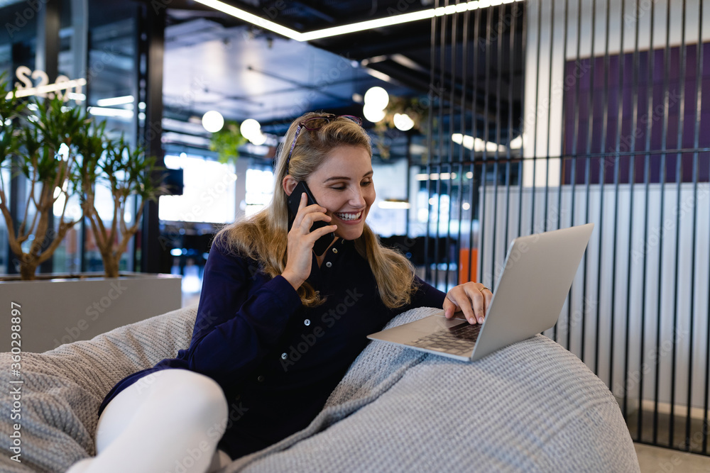 Caucasian woman using a laptop and a phone in a bean bag Caucasian woman using a laptop and a phone in a bean bag