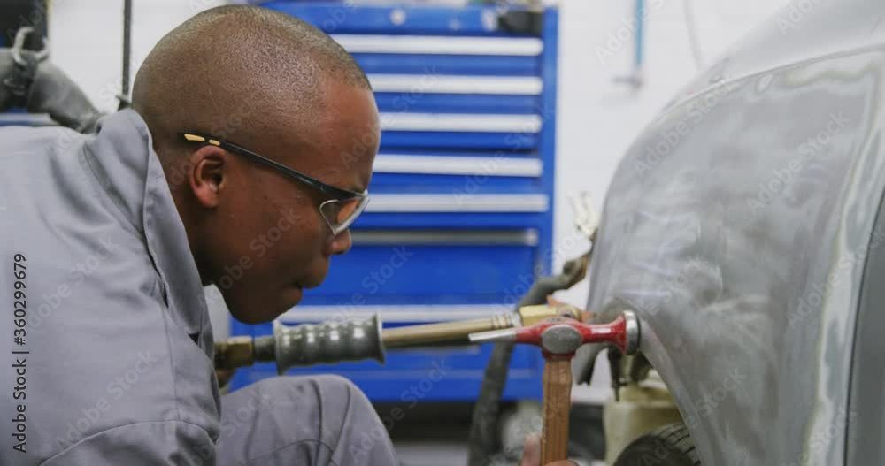 African American male car mechanic holding a screwdriver and using a hammer to repair a car African American male car mechanic holding a screwdriver and using a hammer to repair a car