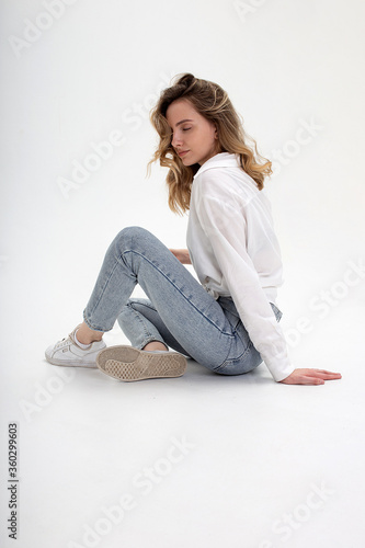 portrait of young caucasian woman posing in shirt and blue jeans, sitting on white studio floor. model tests of pretty girl in basic clothes on cyclorama. attractive female poses with eyes closed