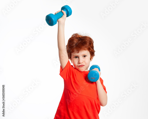 A beautiful red-haired boy plays sports with dumbbells. Fitness, physical education. Little strong man. Healthy physical development of children.