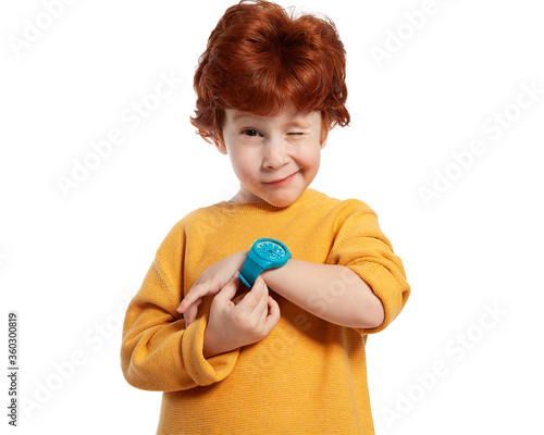 A child with a wristwatch. The red-haired boy shows the time on his watch and squints funny. Portrait of a happy child on a white background. Learning to determine time. Time management.