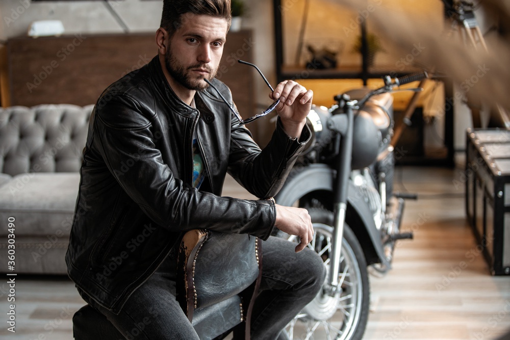 Obraz premium Handsome young man in leather jacket looking away while sitting on the stool