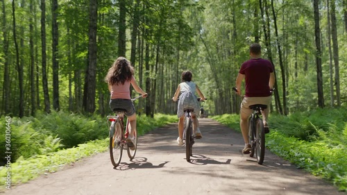 Back view follow shot of happy family of father, mother and little daughter riding bicycles together down trail in green forest