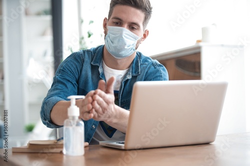 Coronavirus. Man working from home wearing protective mask. Cleaning his hands with sanitizer gel