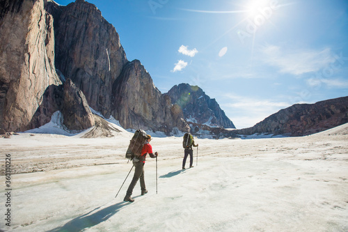 Two backpackers hiking on glacier below steep mountains.