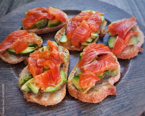 high-quality photo, close-up - on a round dark wooden plate are six delicious juicy crunchy toasts with ripe green avocado and slices of fresh red wild salmon, look appetizing