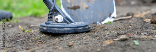 coil electronic metal detector device for search a treasure and the foots of a man on ground background and a shovel next to it. banner