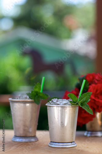Mint Julep drinks in traditional silver cups, vertical, with red roses and horse silhouette in background.