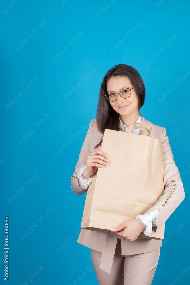 A young pretty girl in glasses and business clothes on a blue background with a paper bag in her hands. A modern woman holds a shopping bag from a store. Concept Of Ecology. Free space. Mock up A young pretty girl in glasses and business clothes on a blue background with a paper bag in her hands. A modern woman holds a shopping bag from a store. Concept Of Ecology. Free space. Mock up