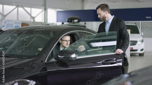 Auto dealer gives key to businessman. Auto dealer opens a new car for auto dealership customer. Businessmen is sitting in a car. Auto dealer shows details to customer.